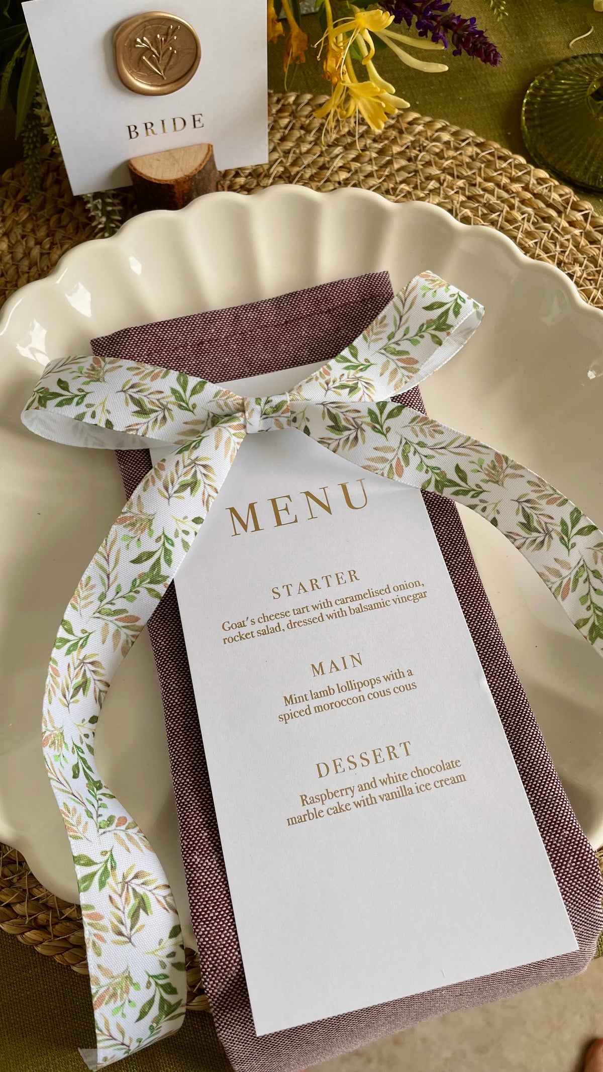 a white rectangle wedding dinner menu card with gold foil text and a botanical ribbon bow laid on top with a wax seal name card for each place setting