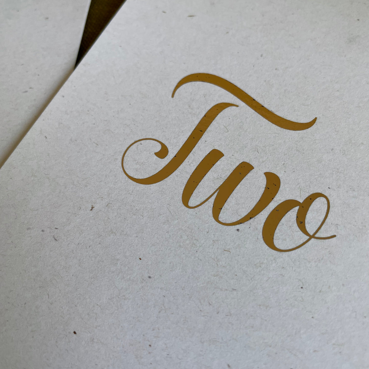 A close-up of a textured off-white paper card featuring the word 'Two' printed in elegant gold cursive script.