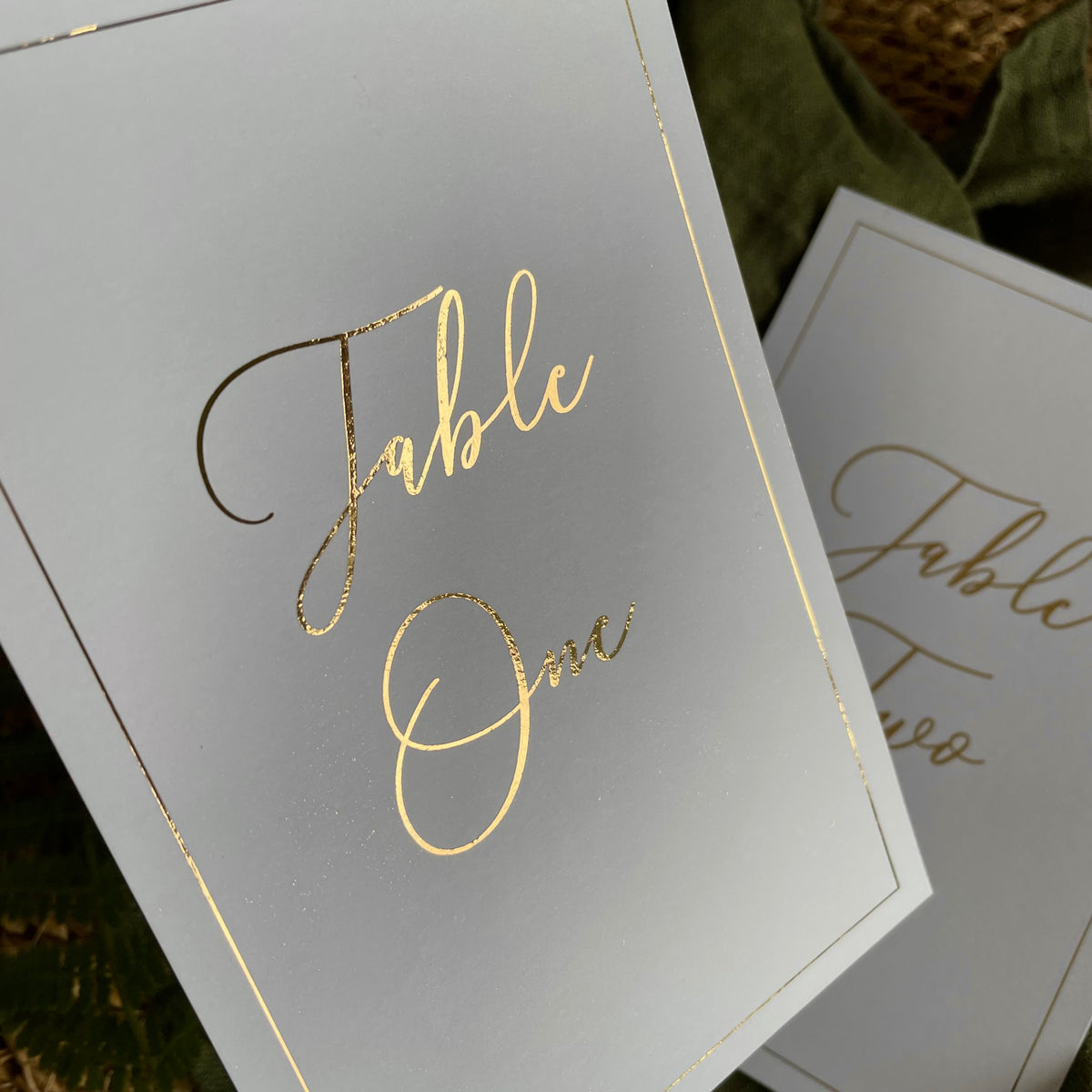 White table number cards with elegant gold foil script reading 'Table One' and thin gold border lines