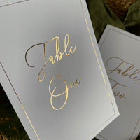 White table number cards with elegant gold foil script reading 'Table One' and thin gold border lines