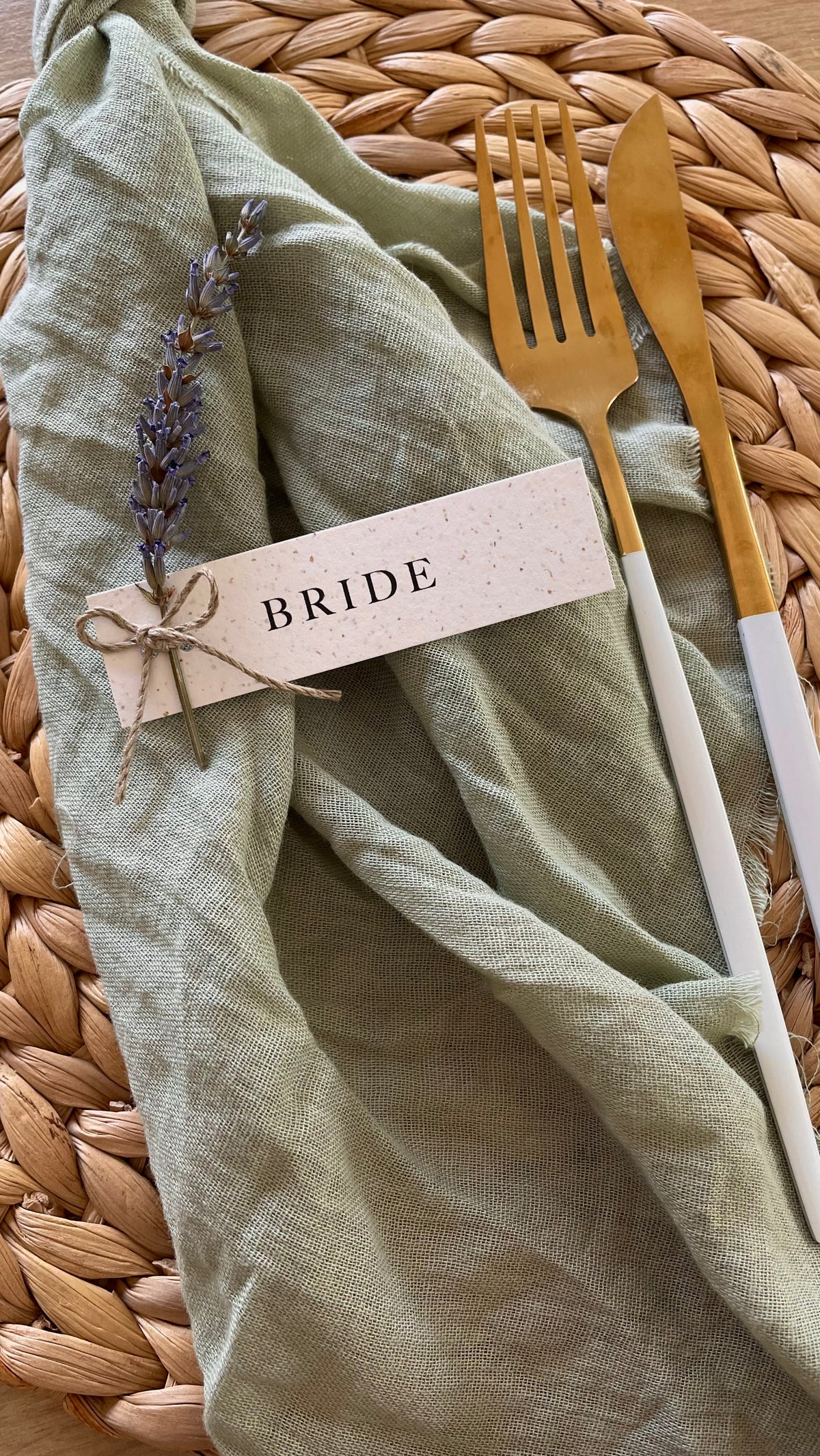 A styled flat-lay of a sage green linen napkin on a woven straw placemat, featuring a gold and white dinner fork, a gold and white knife, and a paper place card tied with twine that reads 'BRIDE' and is decorated with a sprig of lavender.