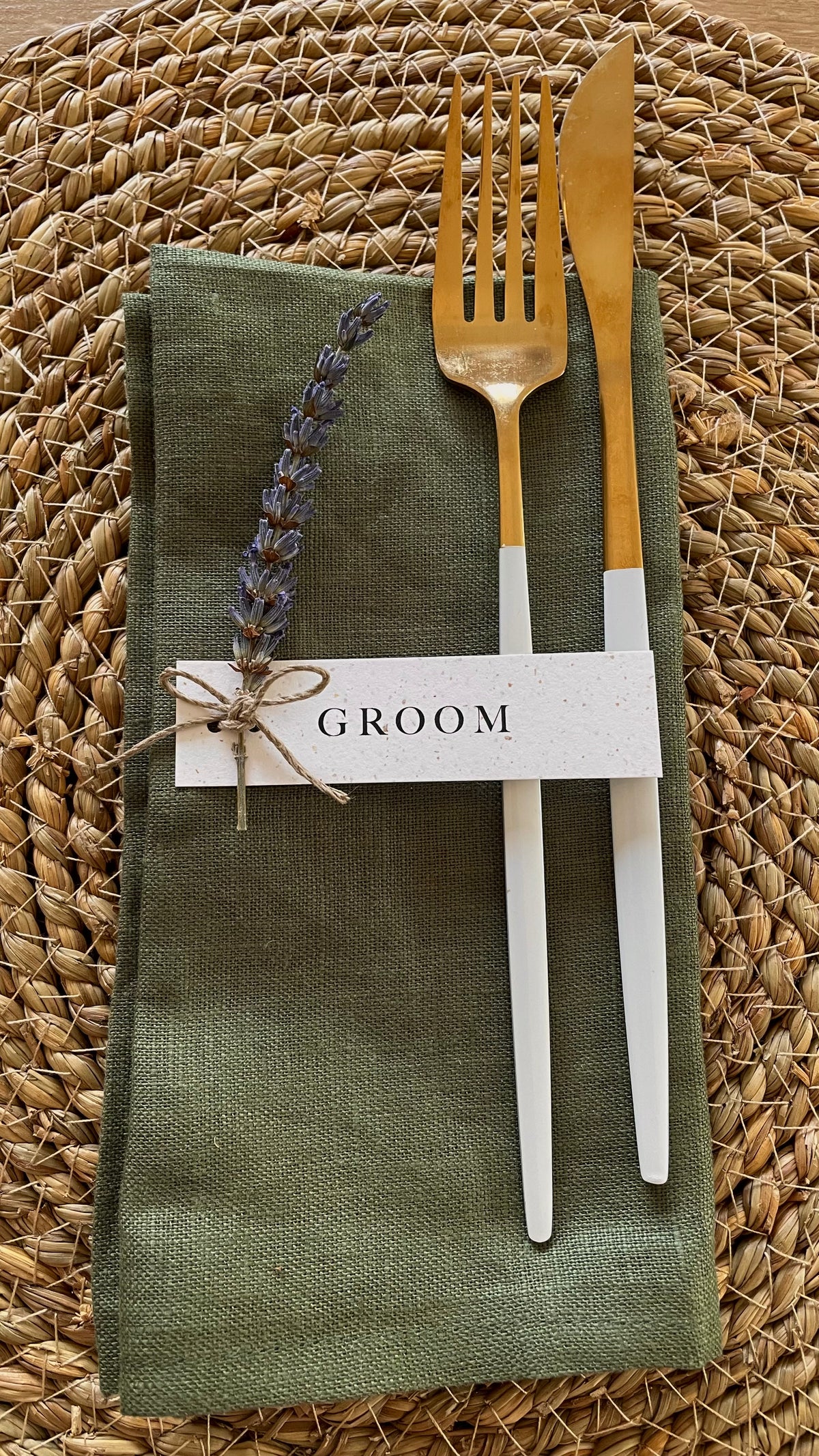 A styled table setting featuring a folded olive green linen napkin on a woven straw placemat, with a gold fork and gold knife, a sprig of lavender, and a white paper tag tied with twine that reads 'GROOM'.