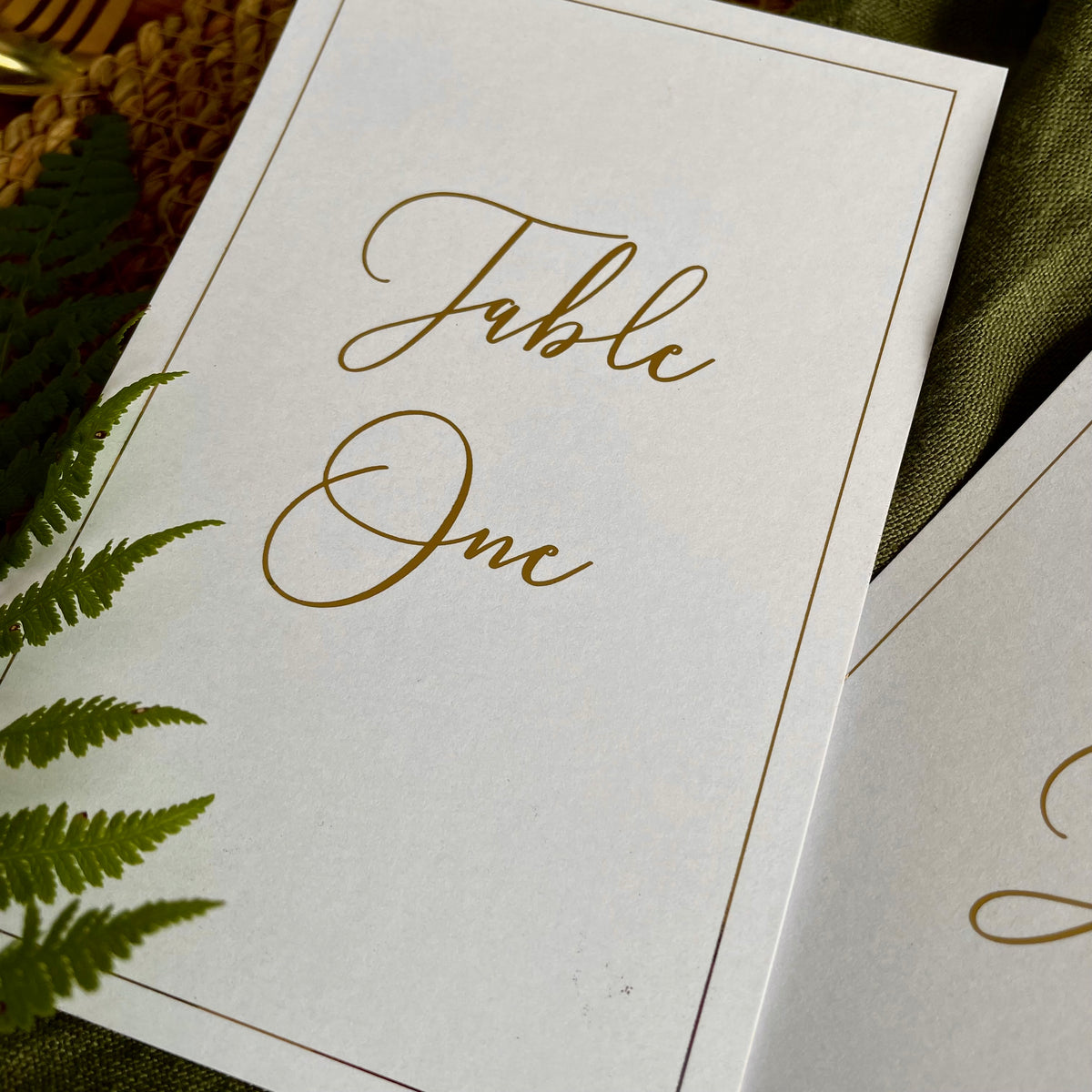 A close-up of a white table number card with elegant gold foil script reading 'Table 1', for rustic elegant wedding reception