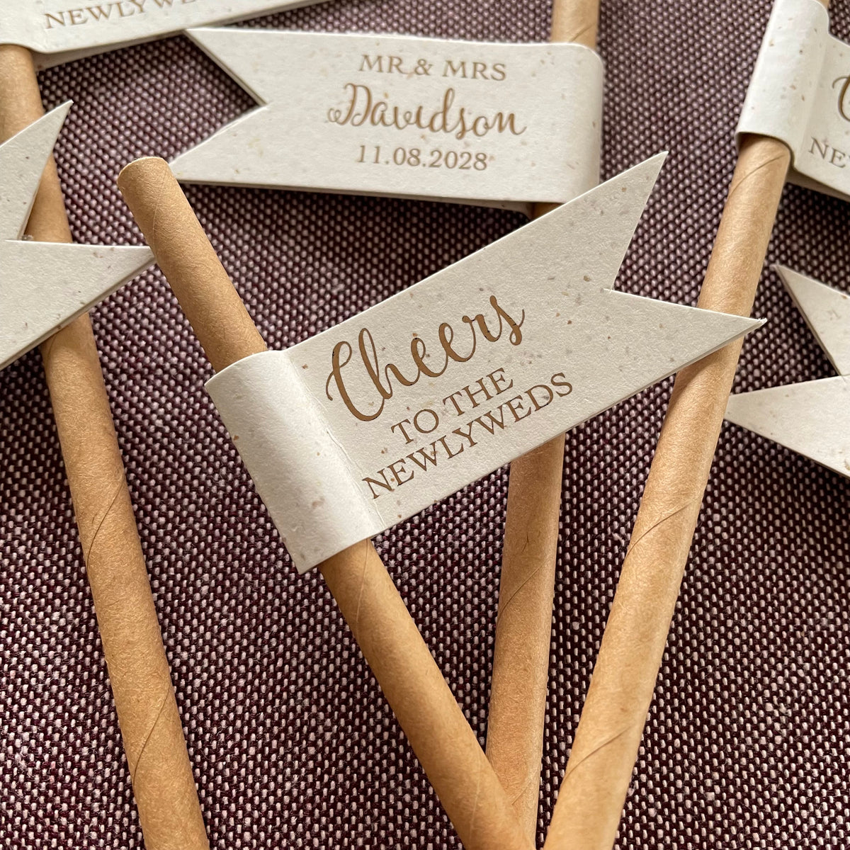 Rustic wedding-themed paper drink straws flags with personalised text on the back. cheers to the newlyweds on the front of the flag in bronze foil.