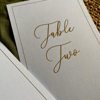 A close-up of a white table card with elegant gold foil script reading 'Table Two,'for elegant rustic wedding table styling