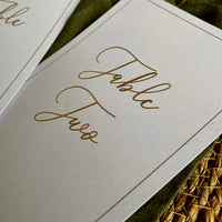 Close-up of a white table number card with elegant gold cursive script reading 'Table Two' for wedding tables